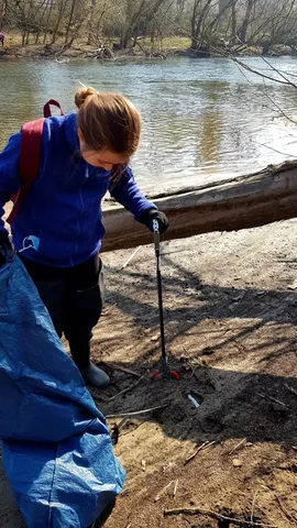 Eine Person sammelt mit einer Müllzange und einem großen blauen Müllsack Abfall am Ufer eines Flusses.