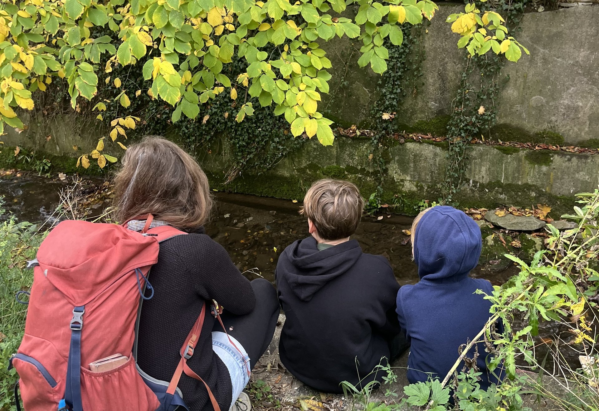Drei Personen, zwei Kinder und eine Begleitperson mit rotem Rucksack, sitzen am Ufer eines kleinen Baches. Über ihnen hängen gelbgrüne Blätter, und im Wasser spiegelt sich das Herbstlicht.