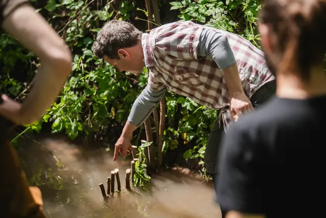 Ein Mann zeigt auf abgesägte Äste am Ufer eines kleinen, bewachsenen Gewässers, umgeben von weiteren Personen.
