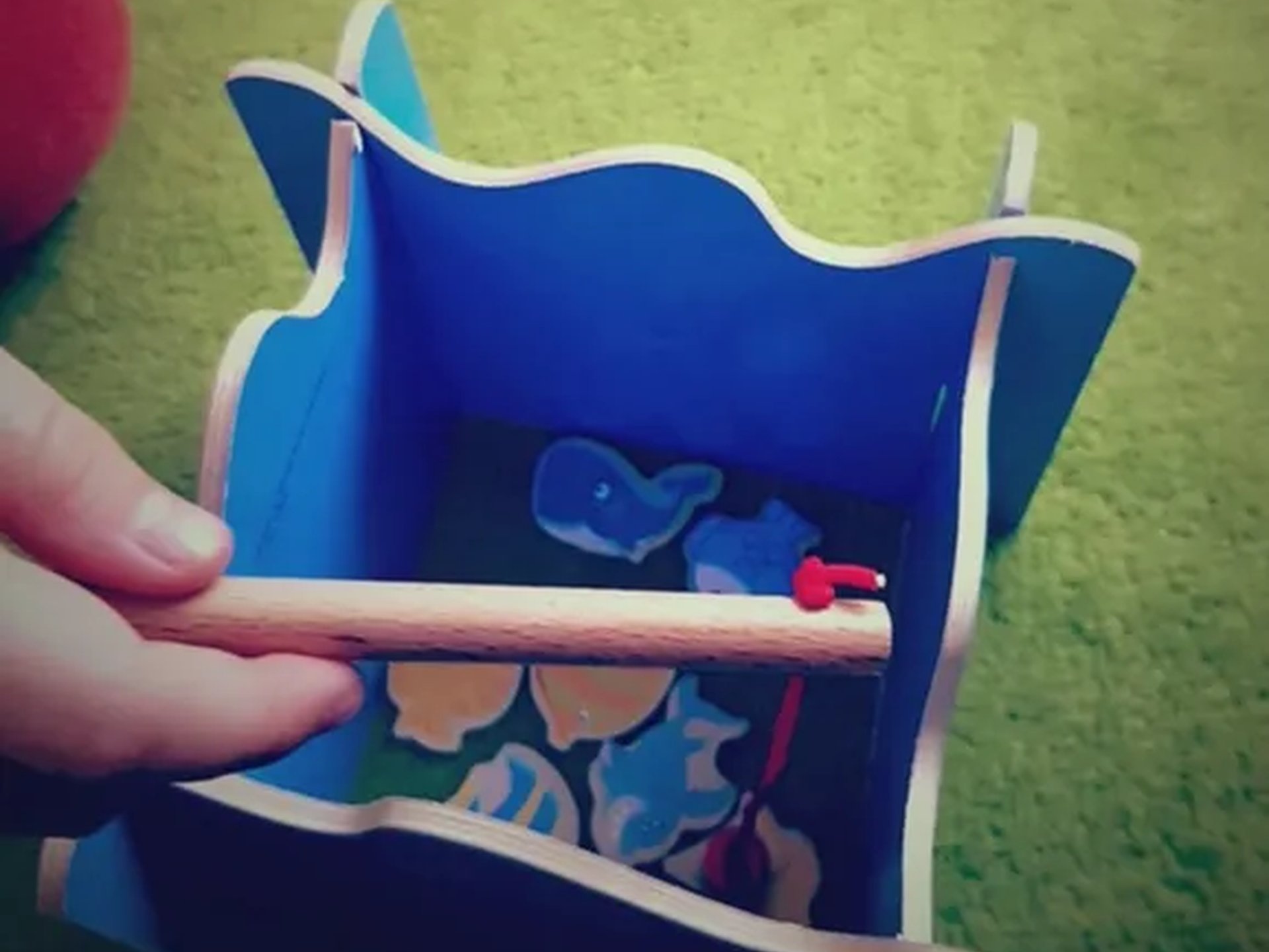 Ein Kind hält eine Angelrute über eine blaue Holzkiste mit kleinen bunten Holztieren darin.