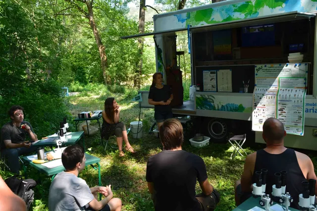 Eine Gruppe von Menschen sitzt im Grünen vor einem mobilen Laborbus und hört einer Person zu, die einen Vortrag hält; auf den Tischen stehen Mikroskope.