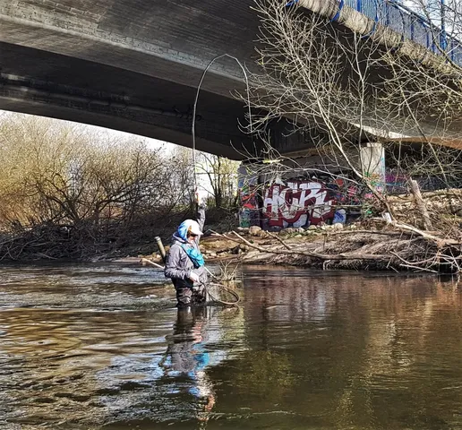 Eine Person angelt im Fluss unter einer Brücke, im Hintergrund sind Graffiti zu sehen.