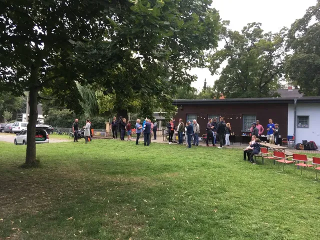 Mehrere Menschen stehen und unterhalten sich vor einem kleinen Gebäude im Grünen; im Vordergrund sind leere Stuhlreihen zu sehen.