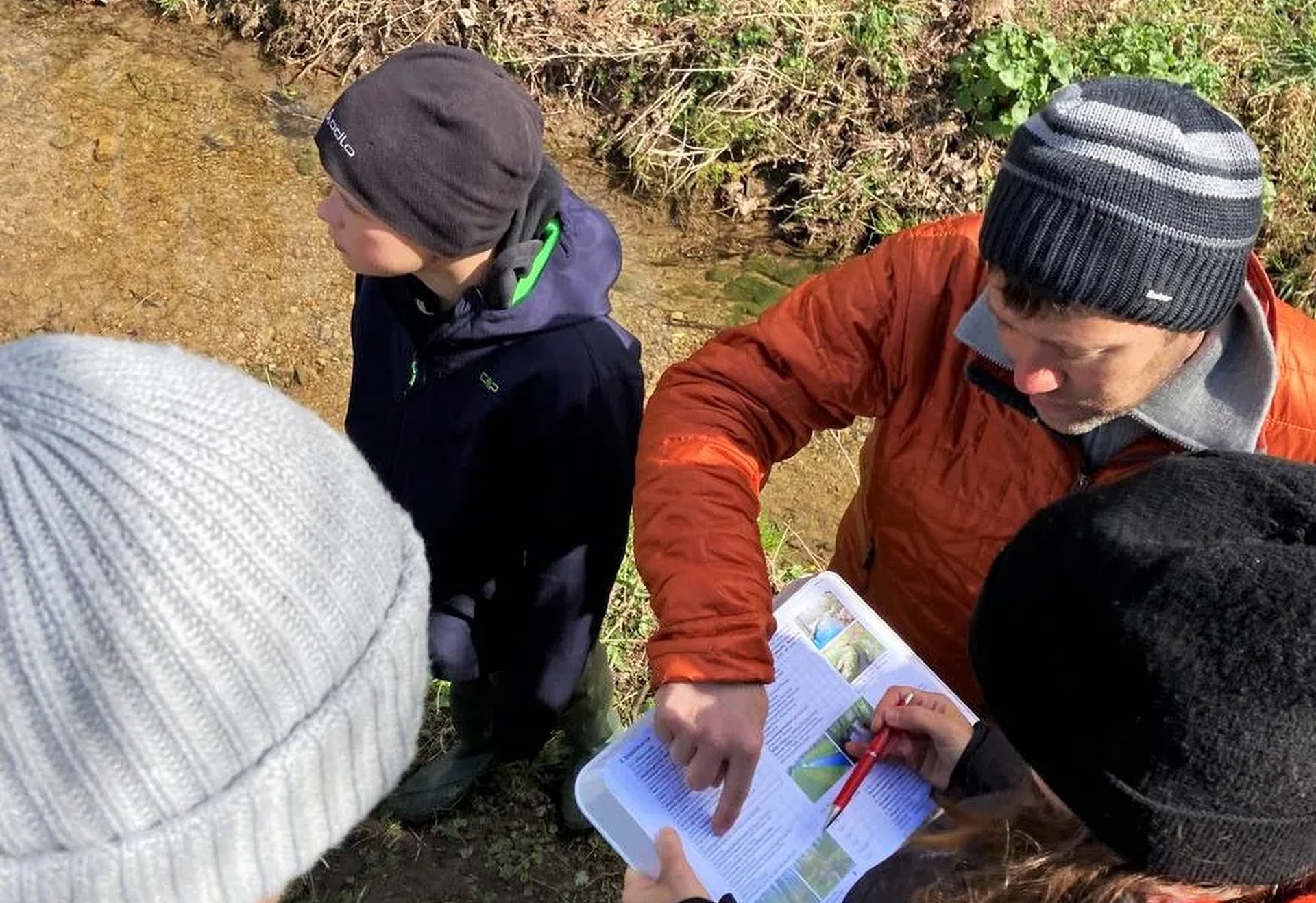 Drei Personen stehen an einem Bach und schauen gemeinsam in ein aufgeschlagenes Buch.