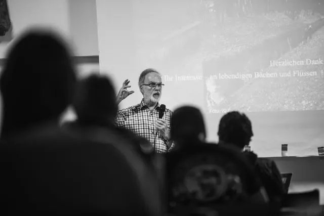 Ein älterer Mann hält einen Vortrag vor einem Publikum, im Hintergrund ist eine Präsentation zu sehen.