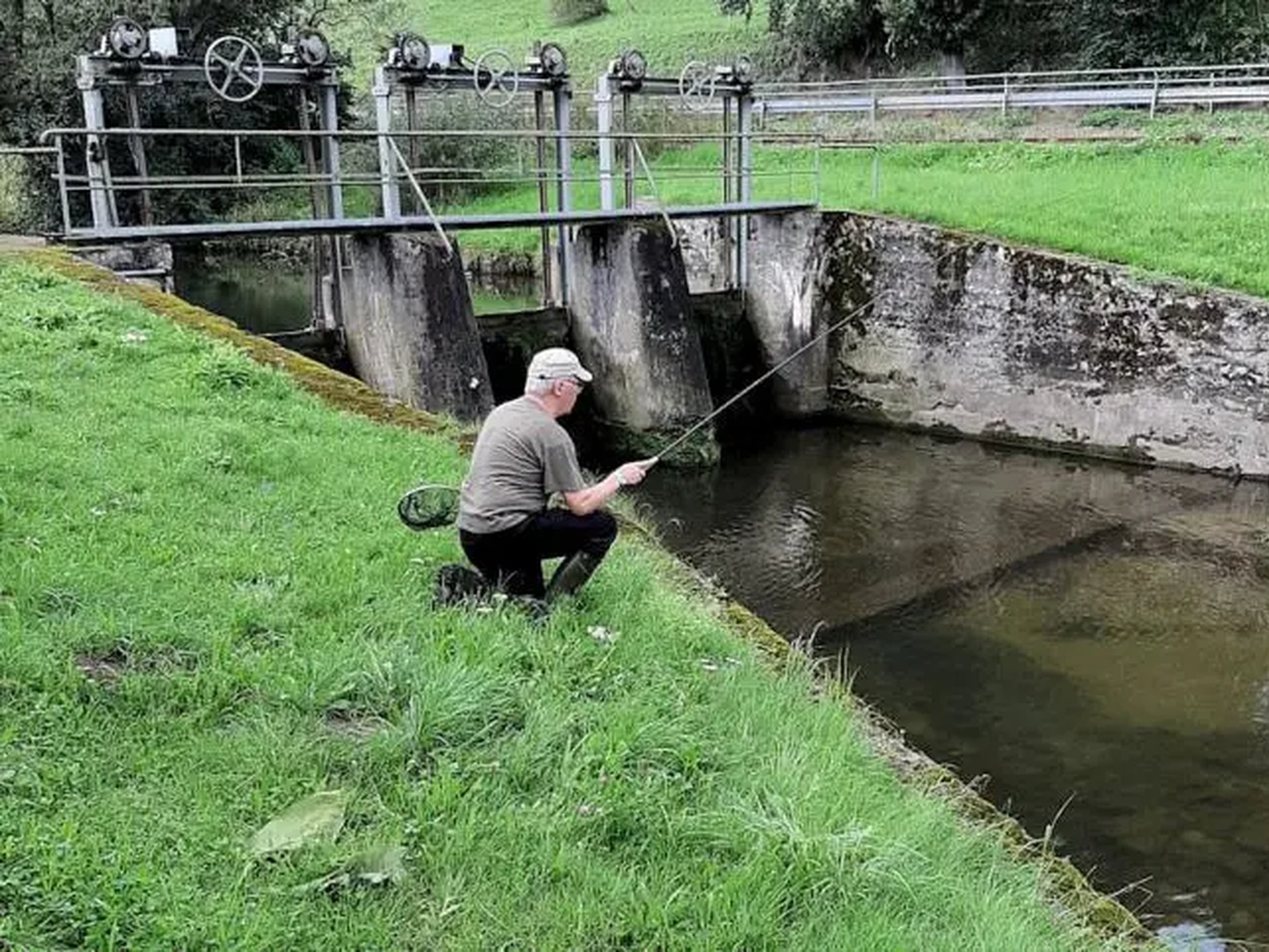 Ein älterer Mann sitzt am Ufer eines kleinen Kanals und angelt mit einer Tenkara-Rute vor einer Stauanlage.