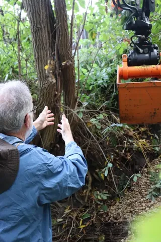 Ein Mann in blauer Jacke steht im Grünen und gestikuliert zu einem orangefarbenen Greifarm einer Maschine.
