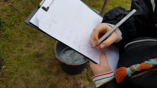 Eine Person hält einen Klemmbrett mit Papier und schreibt darauf, daneben steht ein Eimer mit Wasser auf der Wiese.