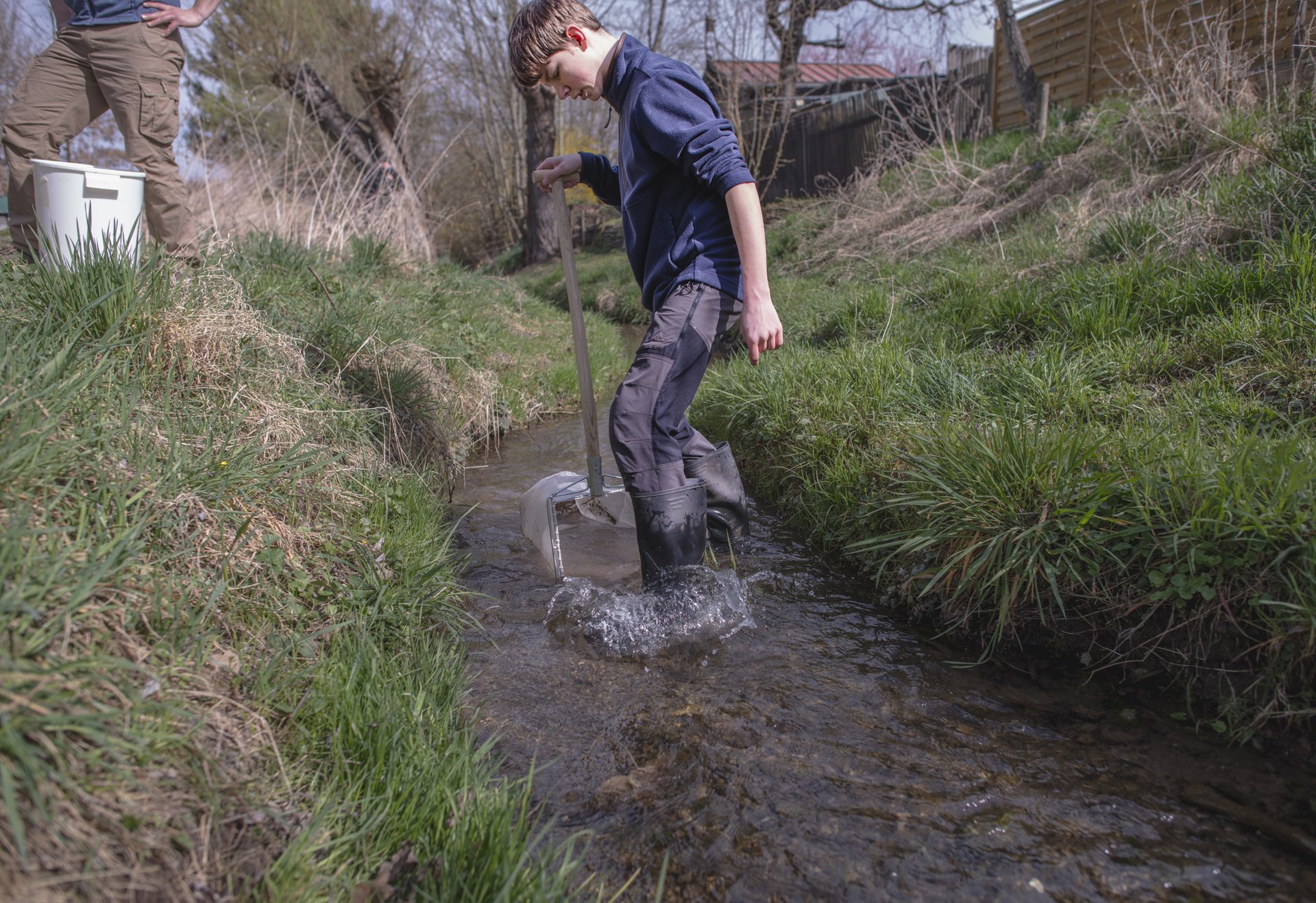 Ein Junge steht mit Gummistiefeln in einem Bach und hält ein Netz im Wasser.