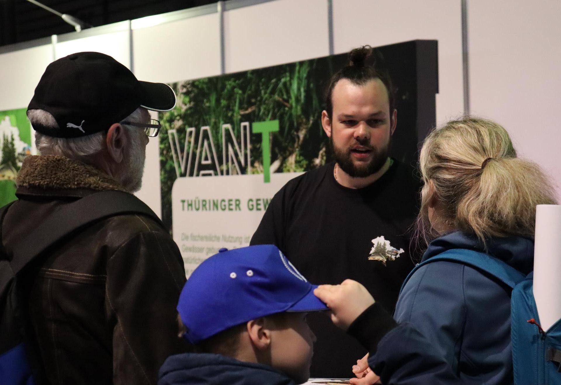 Ein Mann mit Bart spricht an einem Messestand mit drei Besuchern.