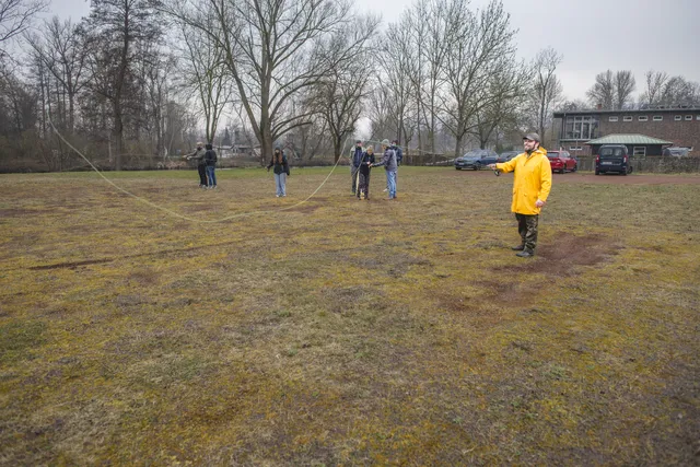 Eine Gruppe Menschen übt das Werfen mit der Fliegenrute auf einer Wiese. Ein Mann in gelber Regenjacke steht vorn und übt eine Wurfbewegung. Im Hintergrund erklärt eine Frau einer Gruppe von Männern etwas.