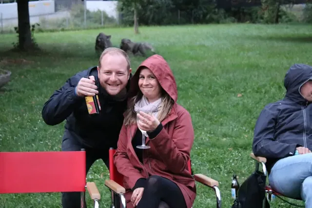 Zwei Personen sitzen auf Gartenstühlen im Freien, beide tragen Jacken, eine Person hält ein Getränk, die andere hält ein Glas; beide lächeln in die Kamera.