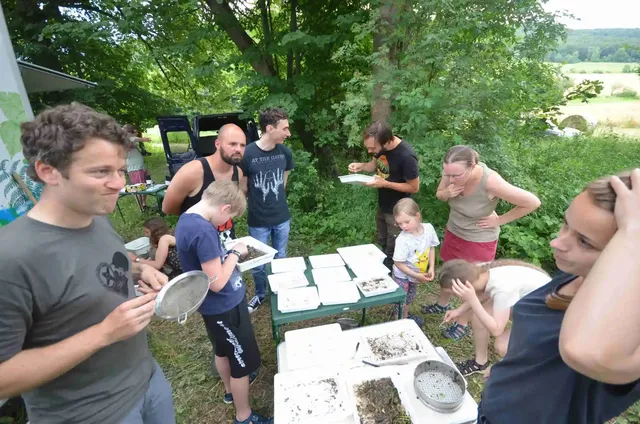 Mehrere Erwachsene und Kinder stehen im Freien um Tische mit Schalen und Untersuchungsmaterial und betrachten interessiert den Inhalt.