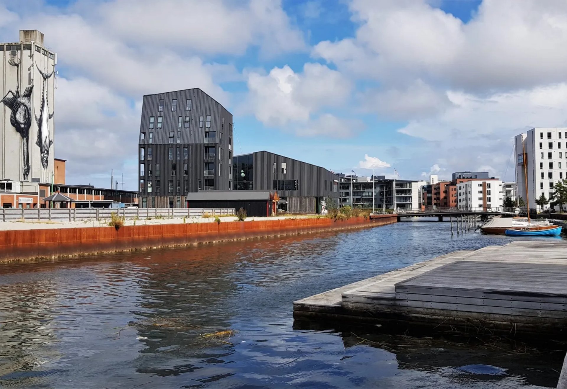 Moderner Stadtteil mit Kanal, Steg und Wohnhäusern; auf einem Turm ist ein großes Wandbild zu sehen.