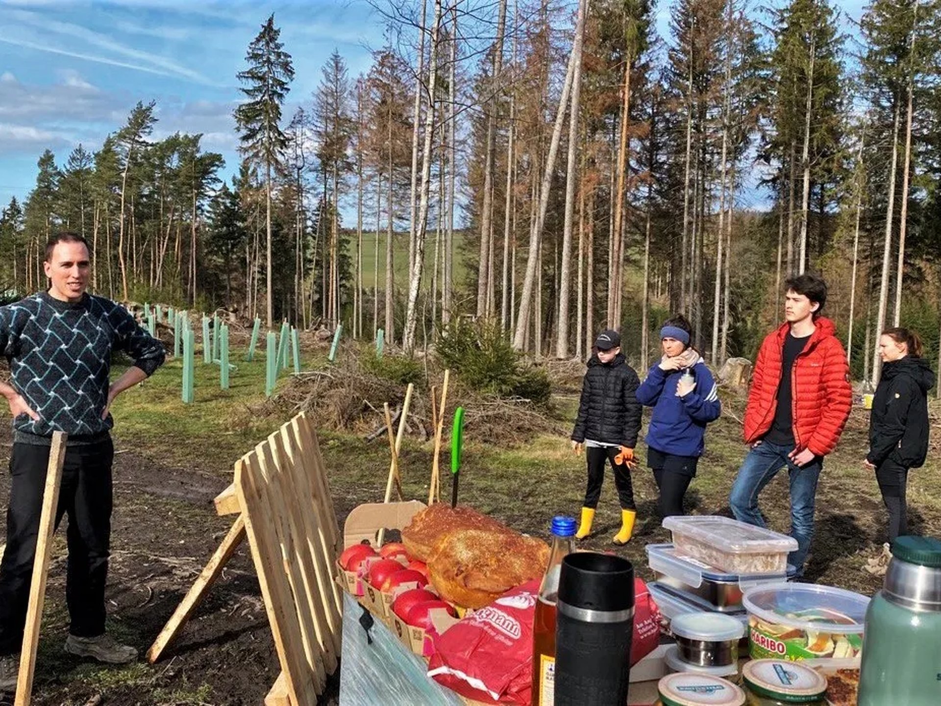 Eine Gruppe von Menschen steht in einem Waldgebiet neben Werkzeugen und einem Tisch mit Verpflegung, während ein Mann spricht.