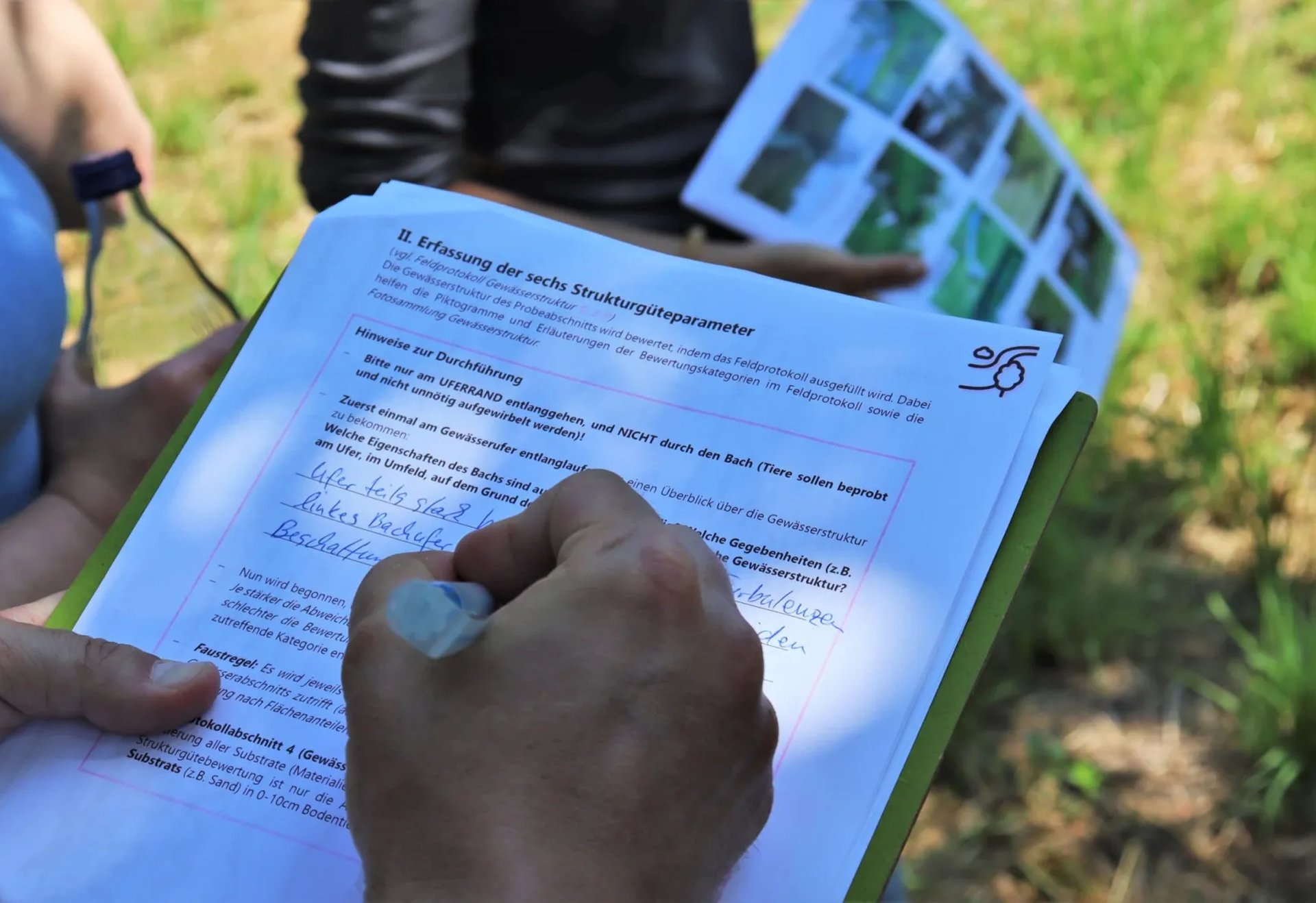 Eine Person füllt im Freien ein Formular auf einem Klemmbrett aus, im Hintergrund hält eine weitere Person ein Blatt mit Bildern.