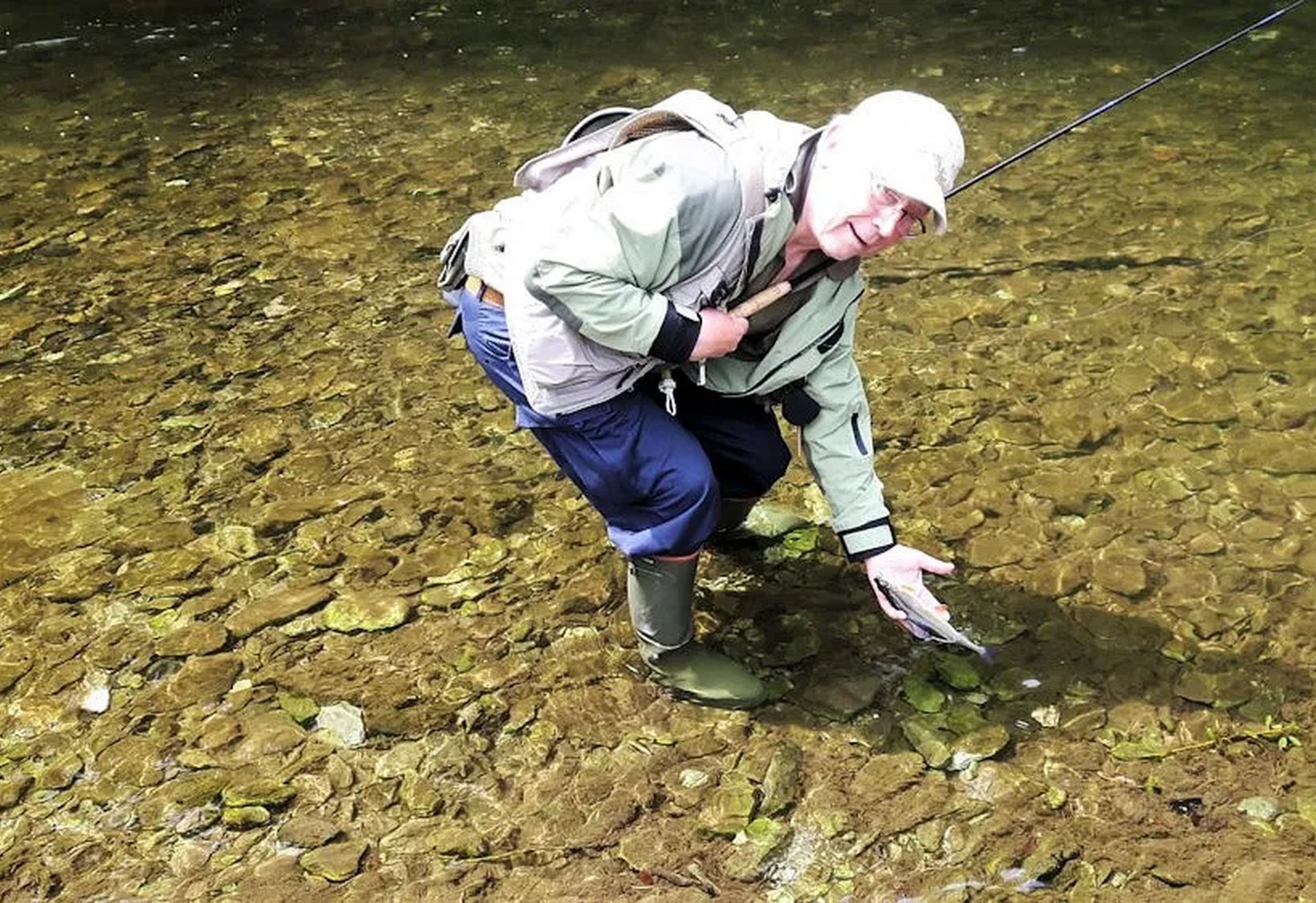 Ein Angler steht in einem klaren Fluss und hält einen gefangenen Fisch im knöchelhohen Wasser.