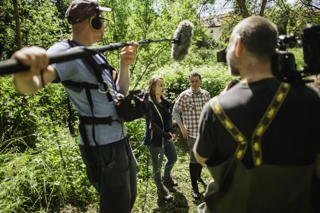 Ein Kamerateam filmt zwei Personen in einem grünen, bewaldeten Außenbereich.