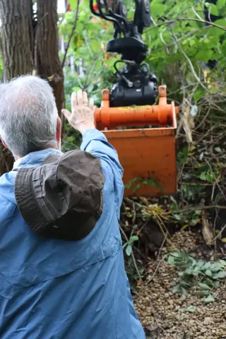 Ein Mann weist einen orangefarbenen Bagger in einem bewaldeten Gebiet ein.