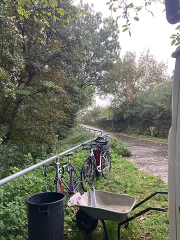 Mehrere Fahrräder stehen an einem Geländer neben einem Weg, davor eine Schubkarre und ein Mülleimer, umgeben von Bäumen und Grün.