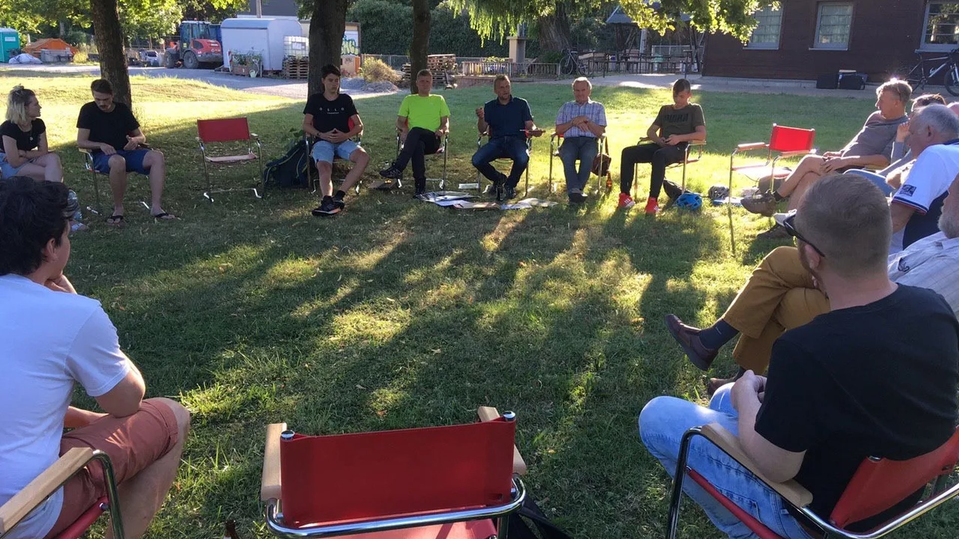 Eine Gruppe von Menschen sitzt im Kreis auf Stühlen im Freien auf einer grünen Wiese unter Bäumen und unterhält sich.