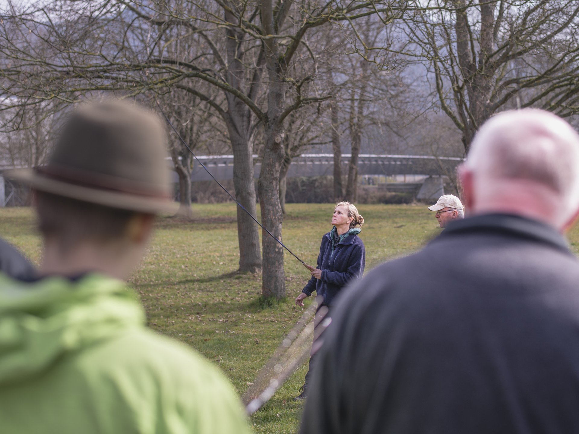 Mehrere Personen stehen auf einer Wiese unter Bäumen, eine Frau in der Mitte übt das Werfen mit der Fliegenrute und schaut dabei der Wurfschnur zu.