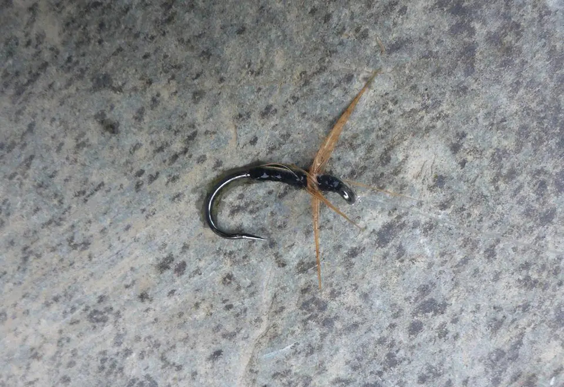 Angelköder mit braunen Federborsten liegt auf grauem Stein.