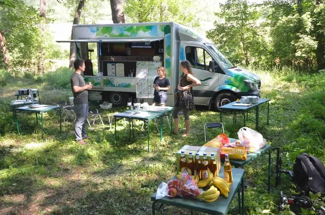 Drei Personen stehen an einem Foodtruck im Grünen, umgeben von Bäumen. Vor dem Truck stehen mehrere Tische mit Lebensmitteln wie Bananen, Getränken und Broten.