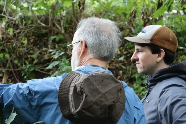 Zwei Männer stehen im Freien vor grünem Gebüsch und schauen in dieselbe Richtung; einer trägt eine Brille und einen Hut am Rücken, der andere eine Basecap.