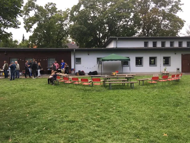 Mehrere rote Stühle stehen in einem Halbkreis auf einer Wiese vor einem Gebäude, in der Mitte befinden sich Tische und ein grünes Partyzelt, im Hintergrund unterhalten sich Menschen in kleinen Gruppen.