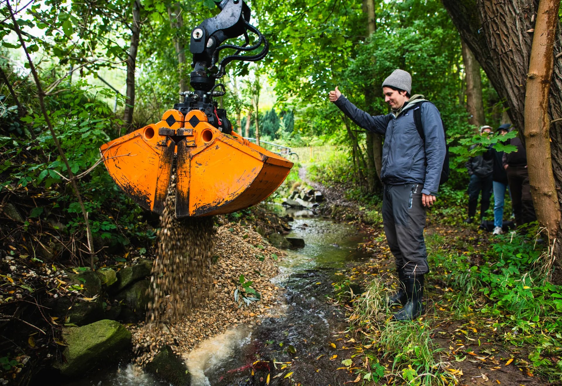 Ein Bagger schüttet Kies in einen kleinen Bach, während ein Mann daneben steht und den Daumen hebt.