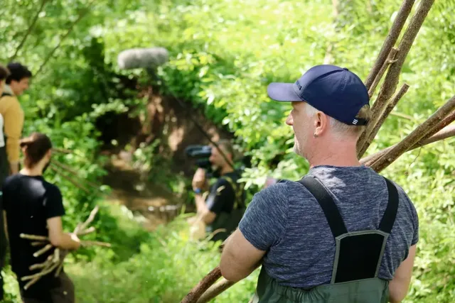 Mehrere Menschen arbeiten im Grünen an einem Bachlauf, einer trägt Äste, im Hintergrund wird gefilmt.