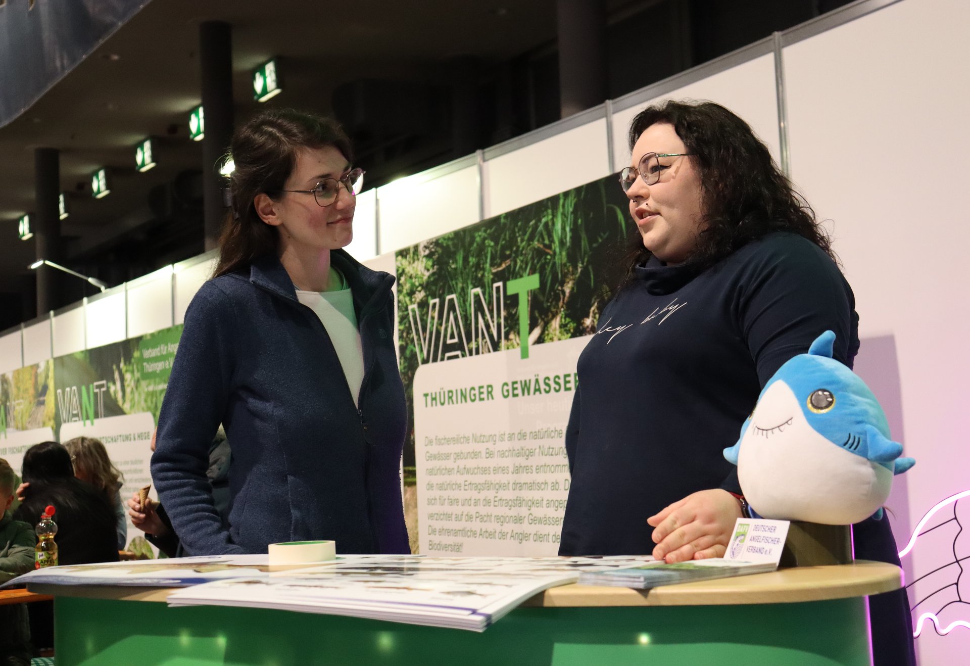 Zwei Frauen unterhalten sich an einem Messestand; auf dem Tresen liegt ein Plüschhai.