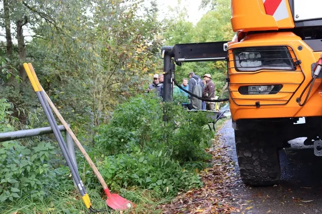 Ein orangefarbenes Baufahrzeug steht am Straßenrand, daneben lehnen Schaufeln und Spaten an einem Geländer; im Hintergrund stehen einige Personen am Straßenrand im Grünen.
