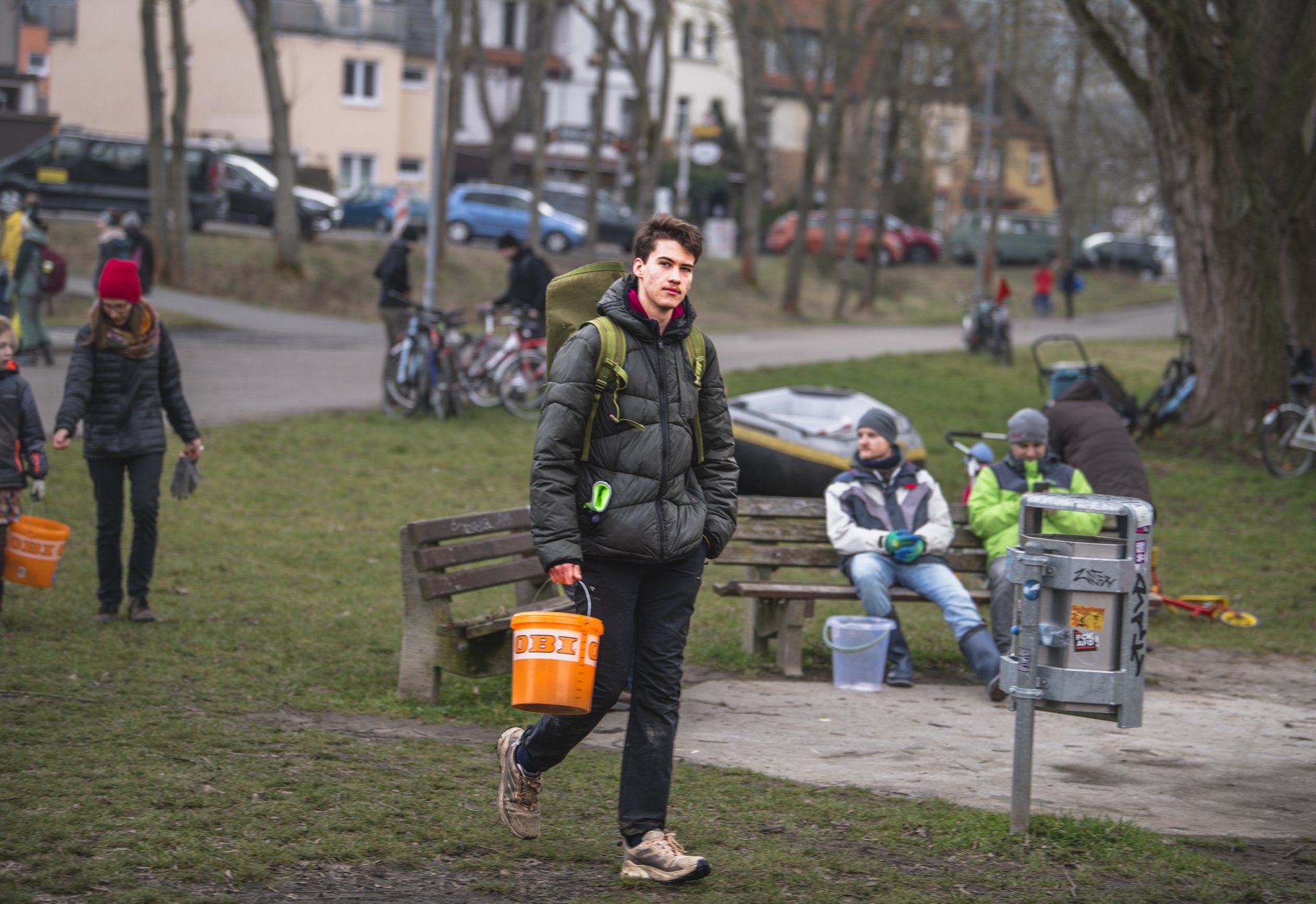 Ein junger Mann trägt einen orangen Eimer in einem Park. Im Hintergrund sind weitere Menschen mit Eimern, Fahrräder und Sitzbänke zu sehen. Die Stimmung ist herbstlich oder winterlich.