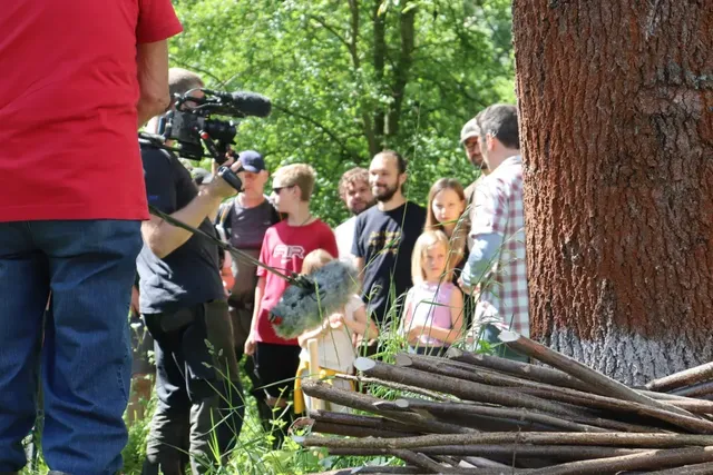Eine Gruppe von Menschen, darunter Kinder und Erwachsene, steht im Grünen neben einem Baum, während ein Kamerateam sie filmt. Im Vordergrund liegen Äste auf dem Boden.