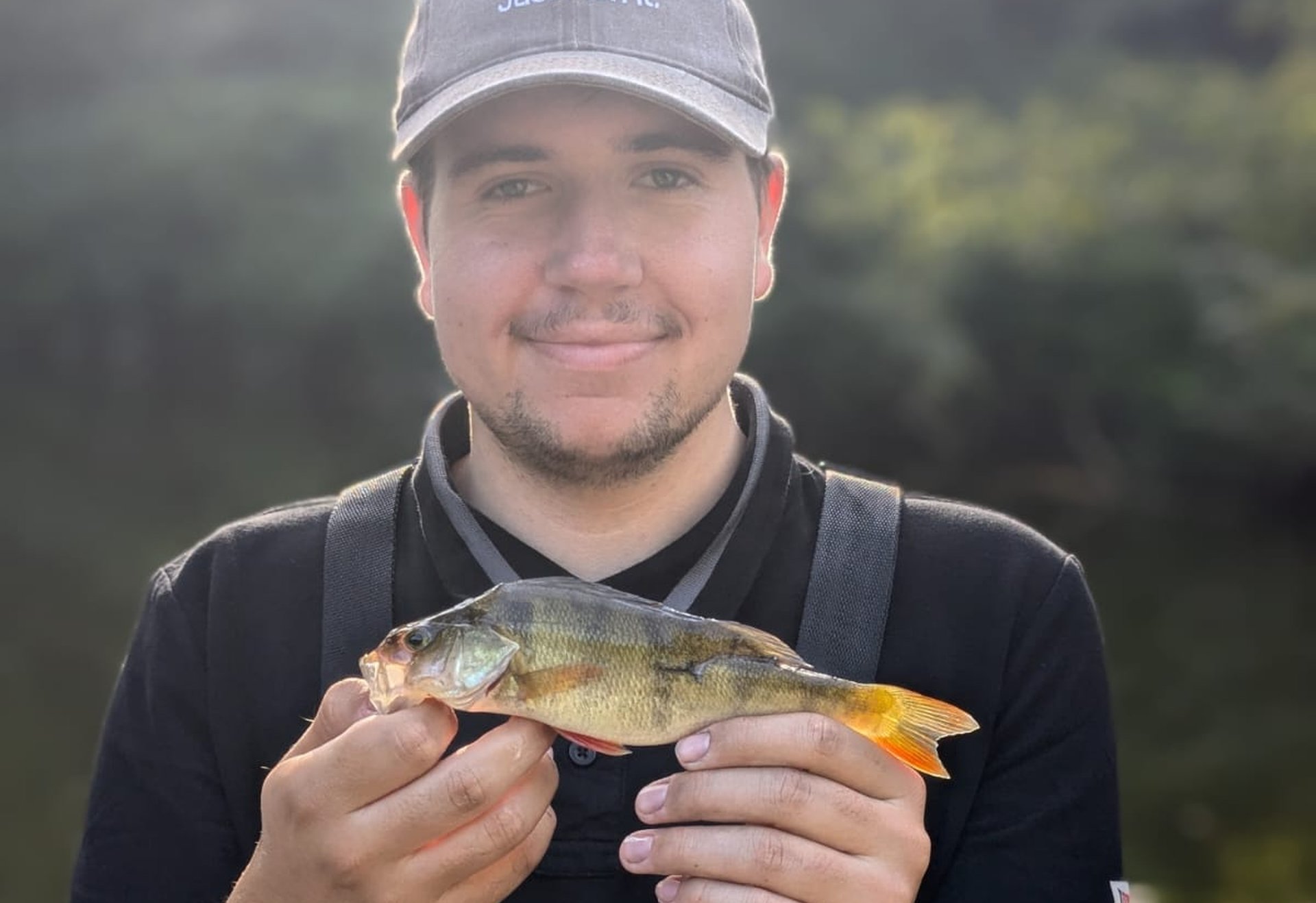 Eine Person mit Kappe hält einen kleinen Barsch vor einem unscharfen Hintergrund aus Wasser und Bäumen.