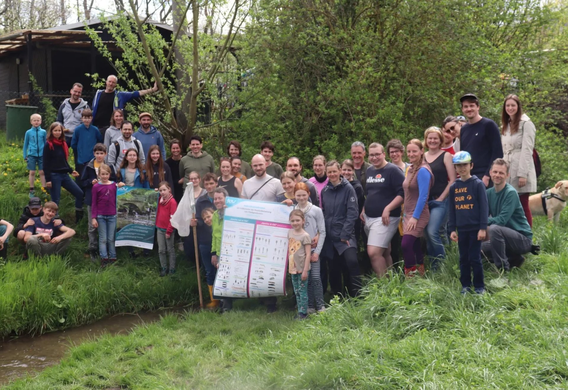 Eine große Gruppe von Menschen steht und sitzt im Grünen an einem Hang neben einem Bach. Einige halten ein großes Plakat, im Hintergrund sind Bäume und eine Holzhütte zu sehen.