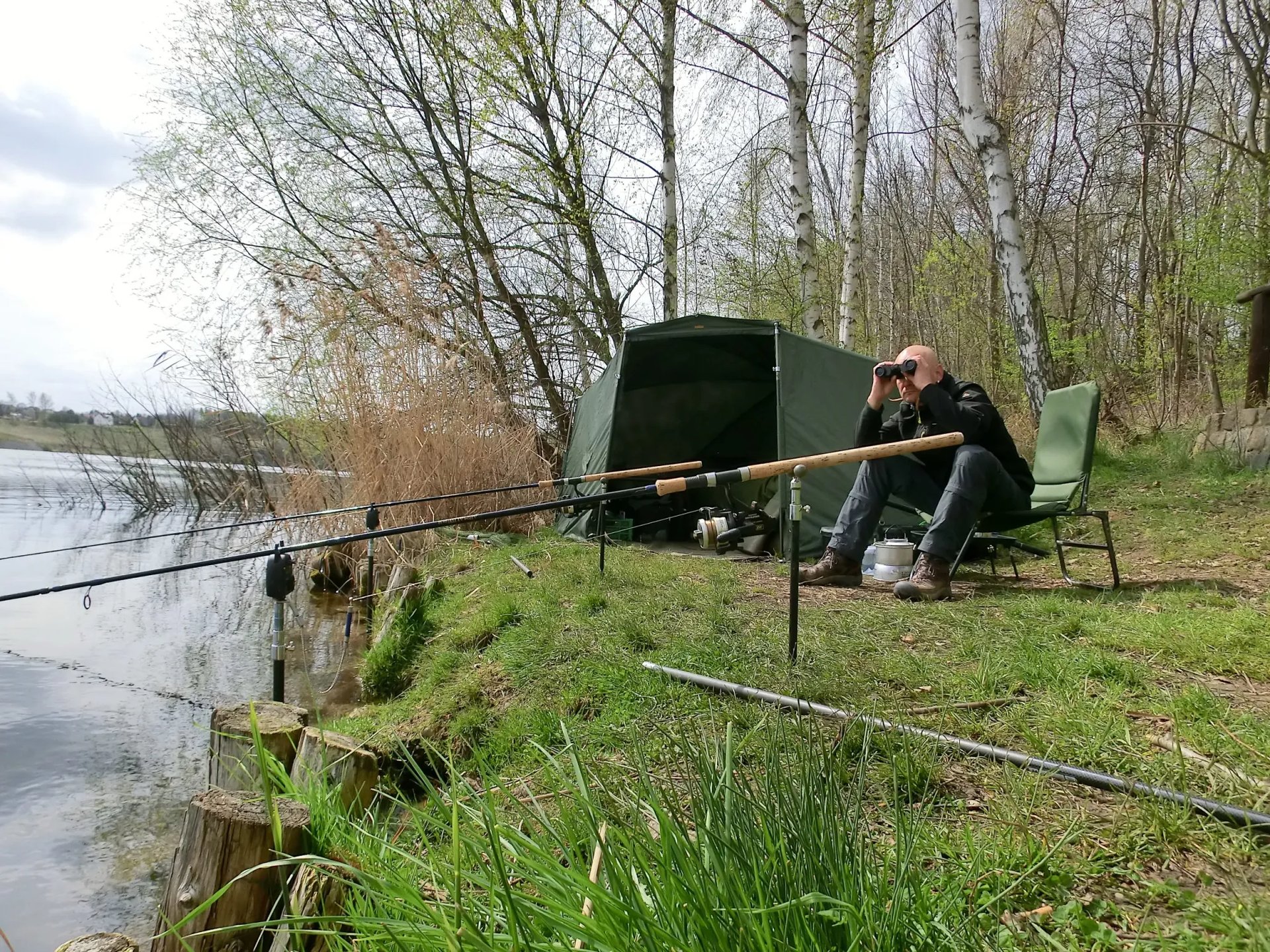 Ein Mann sitzt auf einem Stuhl am Seeufer beim Angeln, mit einer Angelrute vor sich und Fernglas in der Hand, umgeben von Bäumen und einem Zelt.