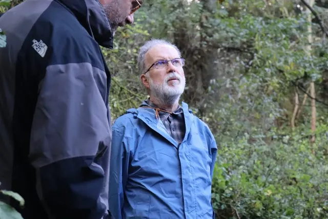 Zwei Männer stehen im Wald, einer trägt eine blaue Jacke und blickt nach oben, der andere eine dunkle Jacke und ist teilweise im Bild.