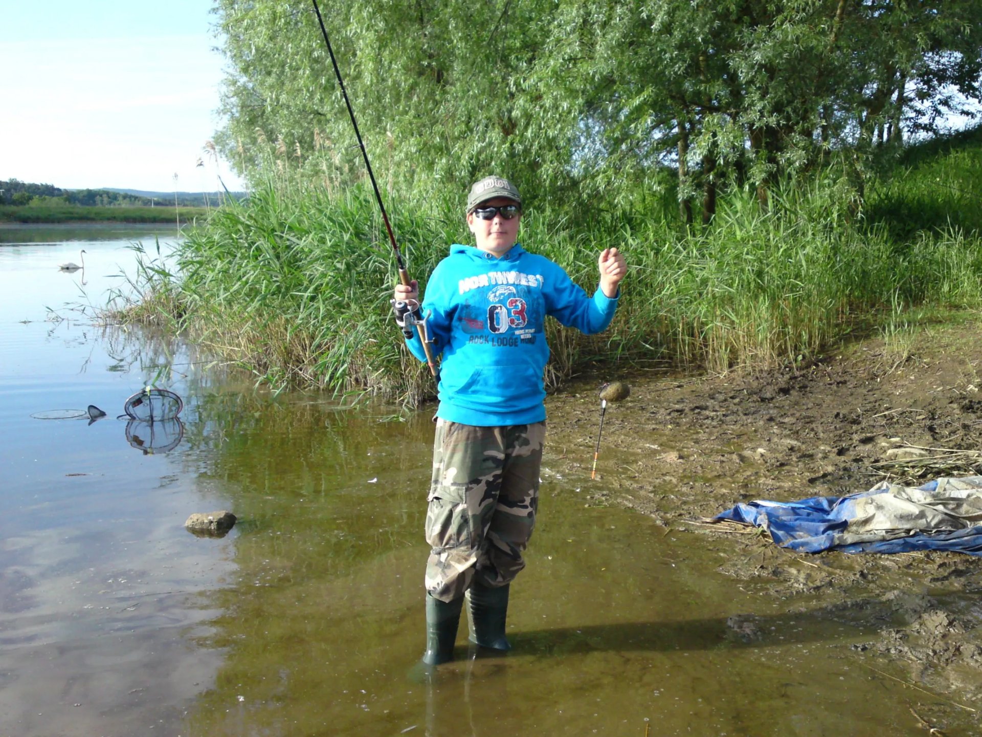 Person in Anglerkleidung steht mit einer Angelrute am Ufer eines Sees.