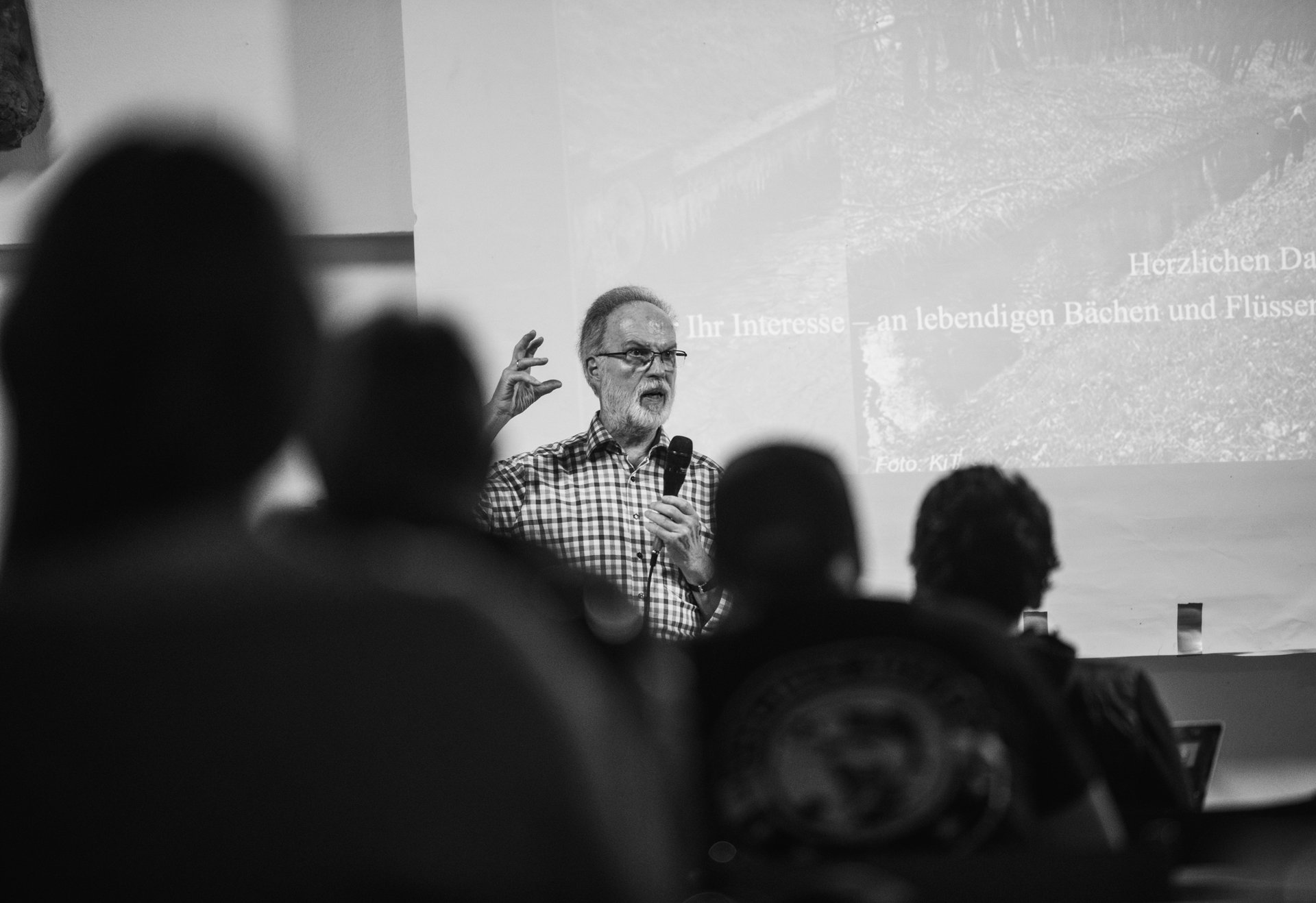 Ein älterer Mann hält einen Vortrag über lebendige Bäche und Flüsse – er spricht mit Mikrofon vor einem projizierten Bild, das Publikum hört aufmerksam zu.