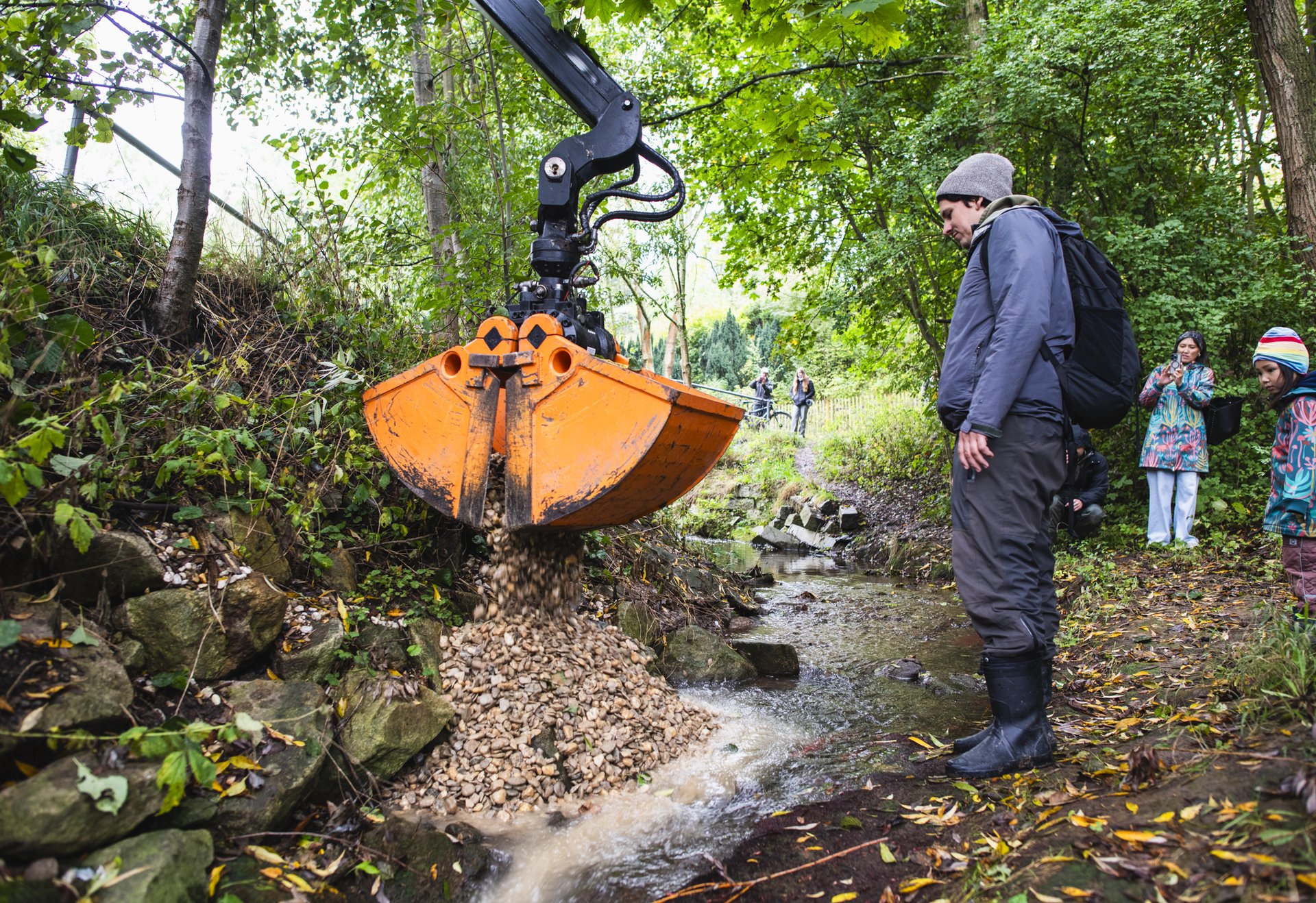 Ein Bagger schüttet Kies in einen kleinen Bach, während mehrere Personen, darunter ein Mann in Outdoor-Kleidung, zuschauen.