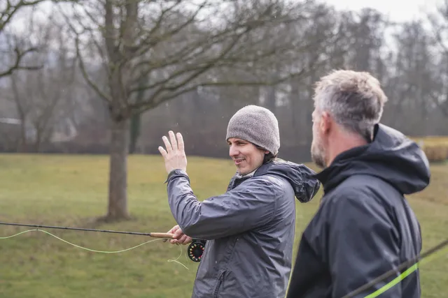 Zwei Männer stehen draußen auf einer Wiese und üben das Fliegenfischen, einer davon trägt eine Mütze und hebt die Hand.