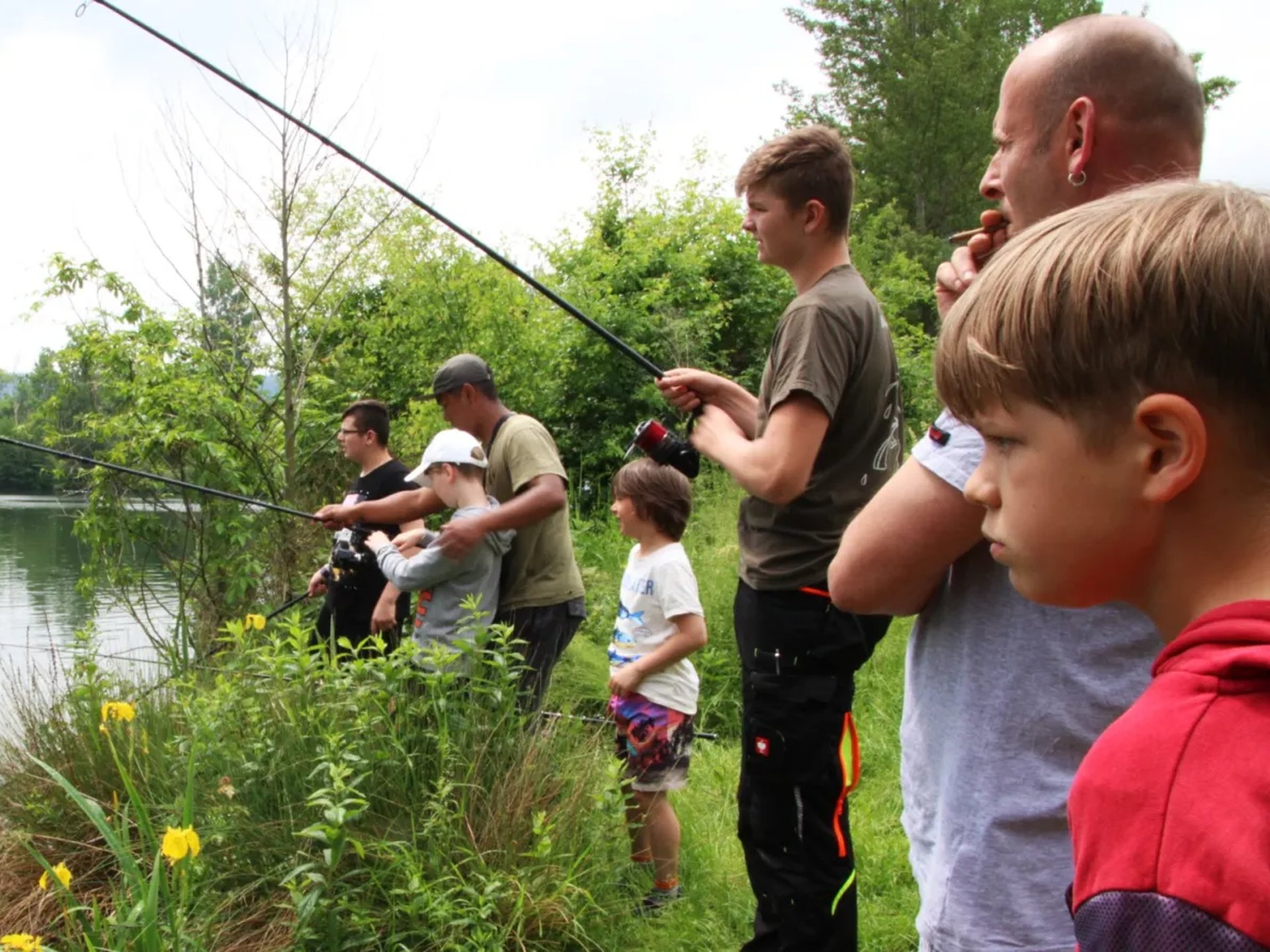 Mehrere Personen, darunter Kinder und Erwachsene, angeln gemeinsam an einem bewachsenen Seeufer.