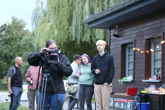 Mehrere Personen stehen im Freien bei einer Holzhütte, im Vordergrund filmt eine Person mit einer Videokamera, im Hintergrund unterhalten sich Menschen; daneben ein Tisch mit Getränken und Snacks.