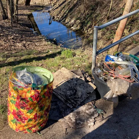 Zwei volle Müllsäcke und Bauschutt liegen neben einem Geländer an einem Bachufer im Freien.