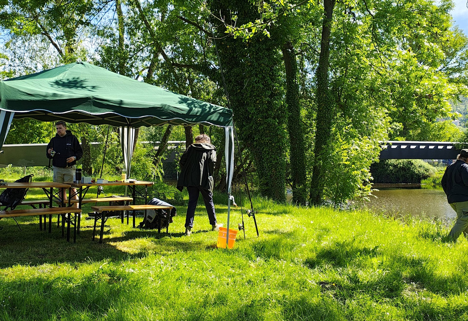 Mehrere Menschen stehen und sitzen unter einem grünen Pavillon an einem Flussufer im Grünen, umgeben von Bäumen und Wiese, im Hintergrund ist eine Brücke zu sehen.