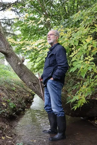 Ein älterer Mann mit grauem Bart steht in Gummistiefeln in einem Bach, umgeben von grünen Bäumen und Sträuchern.