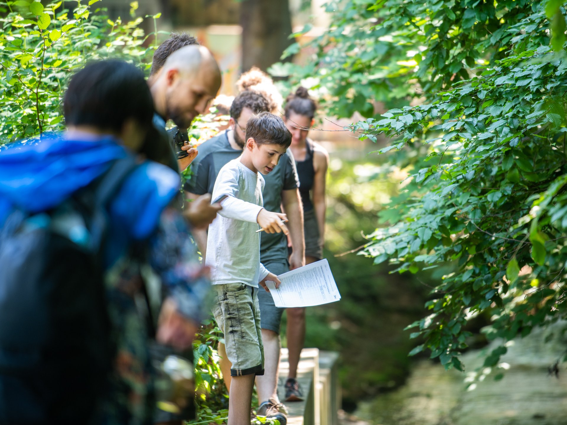 Eine Gruppe von Kindern und Erwachsenen steht an einem kleinen Bach im Grünen. Ein Junge zeigt auf das Wasser und hält ein Blatt Papier in der Hand. Die Szene vermittelt gemeinsames Lernen und Entdecken in der Natur.